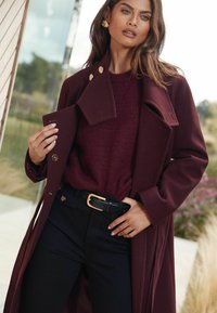 Burgundi gyapjú kabát arany gombokkal egy burgundi kötött pulóver felett. Fekete farmernadrág és fekete öv arany csatot kiegészíti az outfitet.