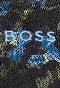Texte "BOSS" brodé en bleu sur un tissu en maille noire avec un fond à motif camouflage bleu, vert et beige.