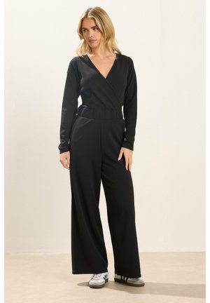 Blondynka ubrana w czarny kombinezon z długim rękawem i dekoltem w kształcie litery V oraz szare trampki, stoi przy jasnej ścianie.