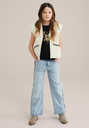 Weißer gesteppter Weste mit schwarzem Besatz, schwarzes Grafik-T-Shirt, hellblaue weite Jeans mit seitlichen Schlitzen und graue ankle boots.