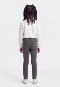 Παιδί με πλεξούδες, φορούν λευκό πουκάμισο μακρυμάνικο, γκρι τζιν και λευκά αθλητικά παπούτσια, στέκεται πλάτη σε απλό φόντο.