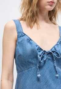 Γυναίκα που φοράει μπλε φόρεμα τζιν χωρίς μανίκια με σούρες στη λαιμόκοψη και φιόγκο μπροστά, απεικονιζόμενη από τους ώμους μέχρι τα μέσα του κορμού.