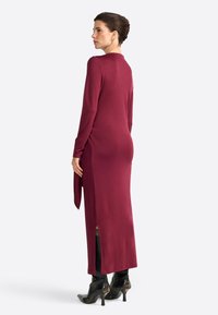 Langes, kastanienbraunes Kleid aus einem weichen Stoff, mit langen Ärmeln und seitlichen Schlitzen. Kombiniert mit schwarzen, hohen Stiefeletten.