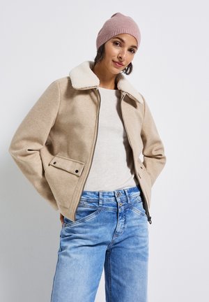 Giacca in misto lana beige con colletto testurizzato color crema, chiusura a zip frontale e tasche laterali. Abbinata a jeans in denim blu chiaro.