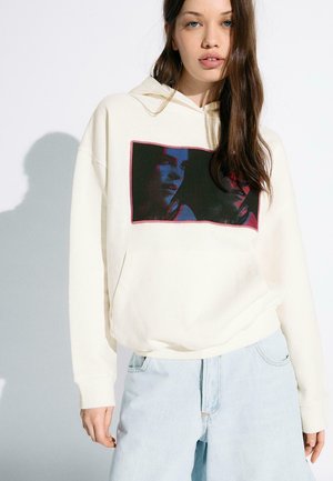 Giovane donna con capelli castani lunghi che indossa una felpa con cappuccio color crema con una stampa scura di un volto e jeans azzurri chiari su sfondo bianco.