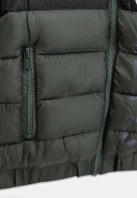 Giacca imbottita verde con materiale elegante, zip verticale e texture trapuntata. Accentuata da un logo discreto e un colore della zip a contrasto.