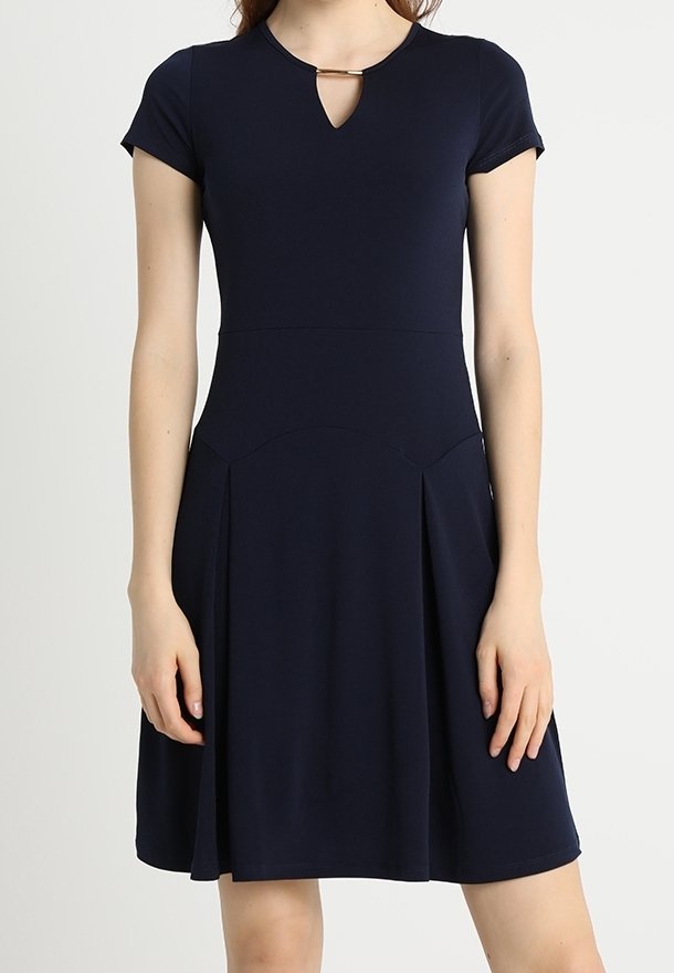 Femme portant une robe bleu marine à manches courtes avec une petite encolure en forme de trou de serrure et un détail barre dorée au col.