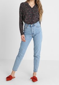 Blouse noire à manches longues avec petit motif floral rouge et blanc, associée à un jean taille haute bleu clair et des sandales à enfiler rouges avec détail nœud.