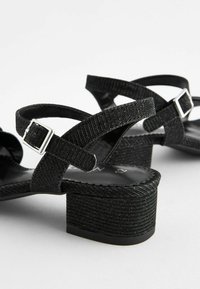 Sorte hælede sandaler har en tekstureret stofoverdel med krydsende remme, justerbare spænder og et chunky hældesign.