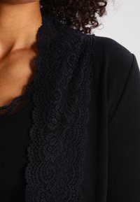Černý top s žebrovanou texturou a širokou krajkovou ozdobou podél výstřihu, s propracovanými vířícími vzory ve tmavém odstínu.