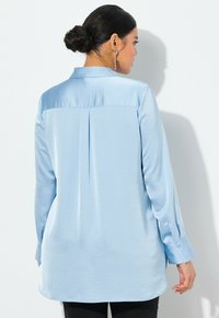 Blusa azzurro chiaro a maniche lunghe realizzata in tessuto morbido. Presenta una piega sul retro, colletto e polsini con bottoni. Design semplice e con una vestibilità rilassata.