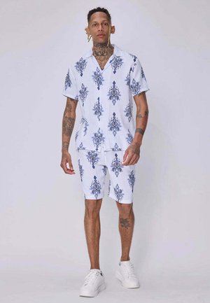 Homme tatoué portant une chemise blanche à manches courtes et un short assortis avec un motif floral bleu et des baskets blanches sur un fond uni.