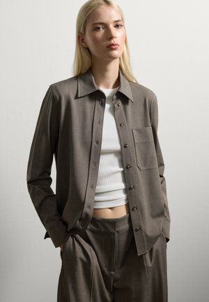 Jeune femme avec de longs cheveux blonds portant une veste marron à boutons, un pantalon assorti et un haut court à côtes blanc contre un mur uni.
