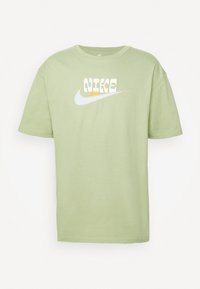 T-shirt vert clair à manches courtes en coton, avec un logo Nike blanc comportant un texte stylisé et un graphique de fleur dans une couleur contrastante.