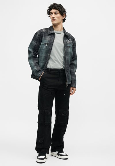 Άνδρας που φορά γκρι μπλουζάκι, μαύρο καρό μπουφάν με φερμουάρ, μαύρο παντελόνι τύπου cargo και ασπρόμαυρα αθλητικά, όρθιος με το ένα χέρι στην τσέπη.