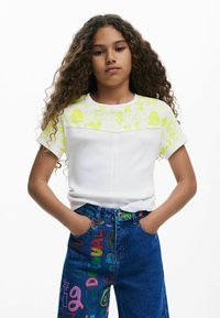 Camiseta blanca con estampados gráficos en neón verde en los hombros; combinada con jeans de mezclilla azul que presentan textos y diseños coloridos.