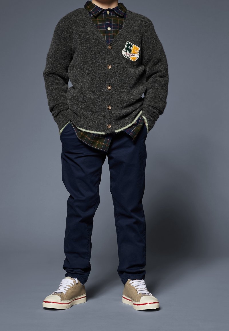 Dziecko w ciemnoszarym kardiganie z naszywką, w kratowaną koszulę, granatowe spodnie i beżowe tenisówki z białymi sznurówkami stoi z rękami w kieszeniach.