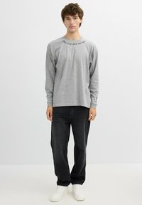 Una camiseta gris de manga larga presenta una frase impresa cerca del cuello, combinada con unos jeans negros y zapatillas blancas, sobre un fondo liso.