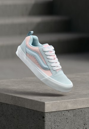 Baskets basses bleu clair et rose avec des lacets et une semelle blanches, en lévitation au-dessus d'un rebord en béton avec des escaliers en arrière-plan.