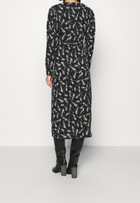 Robe noire à manches longues avec un motif floral blanc, taille cintrée et longueur mi-mollet. Portée avec des bottes noires ajustées jusqu'au genou.
