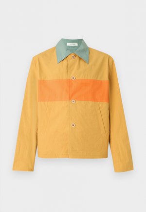 Vīrietis, kurš valkā sinepju dzeltenu jaku ar zaļu apkakli pār melni baltu rūtainu kreklu, melnas platajām biksēm un tumšiem apaviem.