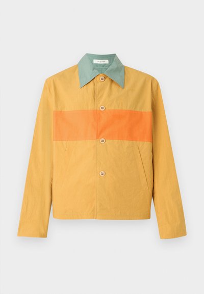 Κίτρινο τζάκετ με φαρδιά πράσινη γιακά, οριζόντια πορτοκαλί ρίγα στο στήθος, κλείσιμο με κουμπιά μπροστά και πλαϊνές τσέπες σε λευκό φόντο.