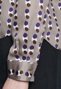 Silkesblus i ljus taupe med abstrakta prickiga mönster i lila, brunt och vitt. Har en knäppt manschett och en mjuk textur.