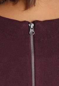 Top de punto burdeos con un cierre de cremallera de metal plateado centrado en la parte posterior. El escote es ancho y acanalado, con una textura suave.