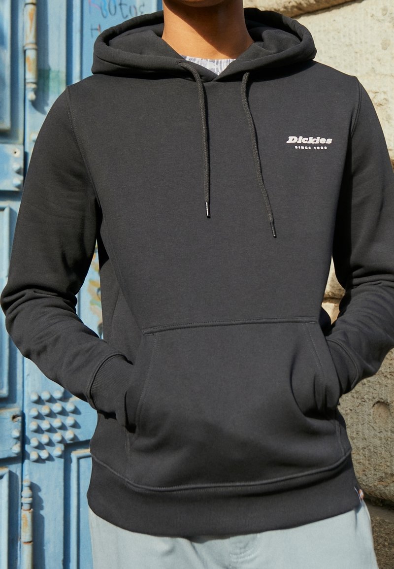 Felpa nera con cappuccio, tasca frontale, cappuccio regolabile con lacci e logo bianco "Dickies". Il tessuto sembra morbido e liscio.