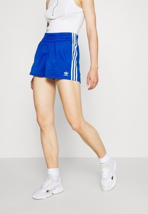Nainen, jolla on päällään siniset Adidas-shortsit valkoisilla raidoilla ja valkoiset lenkkarit, kävelee vaaleaa yksiväristä taustaa vasten.