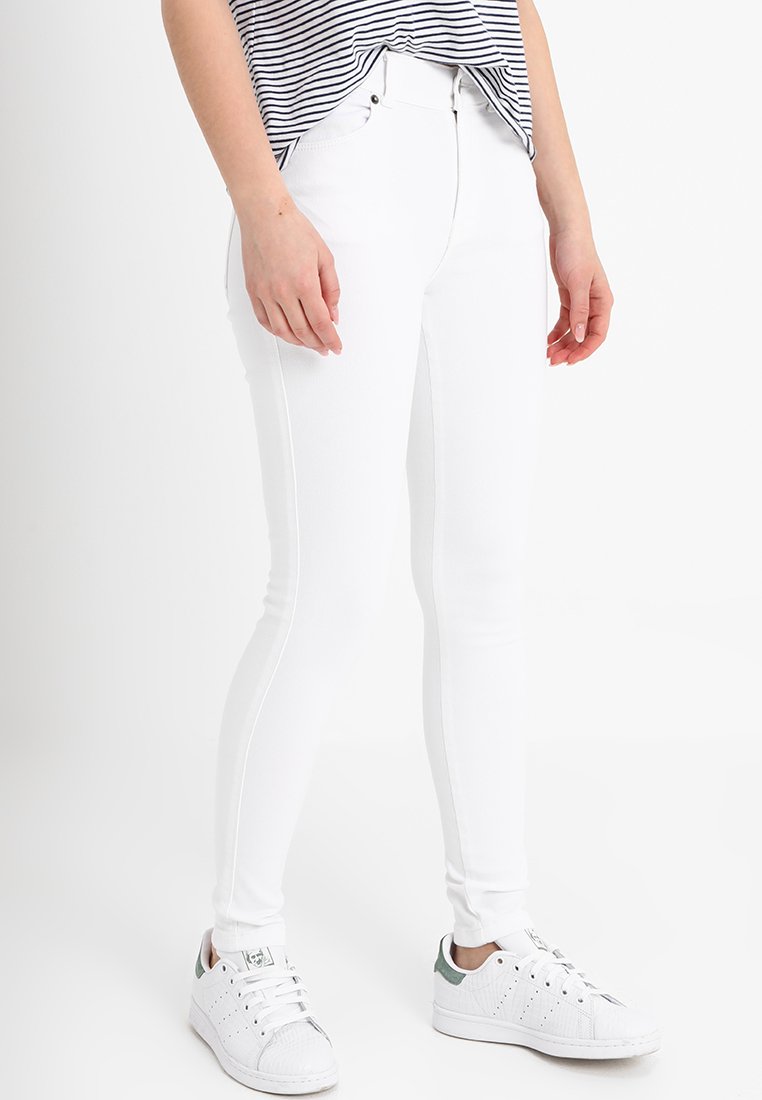 Jeans brancos skinny feitos de um tecido elástico, com costuras laterais subtis. Combinados com tênis brancos com detalhes verdes.