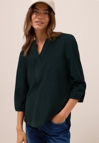 Blusa leggera di un verde scuro con scollatura a V e maniche 3/4. Il tessuto ha una texture liscia. Abbinata a jeans in denim blu.