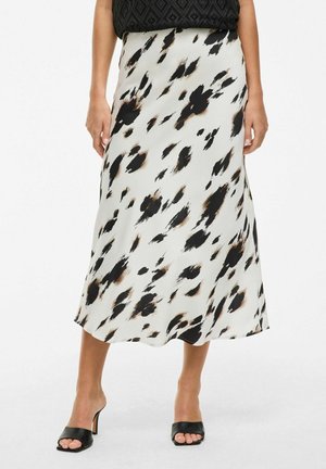 Vrouw draagt een witte midirok met zwarte en bruine abstracte vlekken, gecombineerd met zwarte open teen hoge hak sandalen.