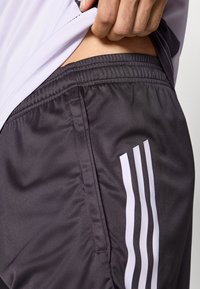 Sivé atletické šortky s elastickým pásom, so tromi bielymi pruhmi po boku a hladkým, lesklým povrchom.