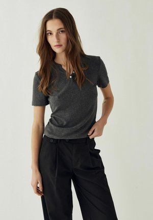 Mladá žena s dlhými hnedými vlasmi, oblečená v tmavosivom tričku s krátkym rukávom a čiernych vysokých nohaviciach, stojaca pred jednoduchým svetlým pozadím.