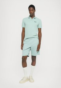 Camisola polo azul clara com colarinho e logótipo EA7, combinada com calções a condizer. O modelo usa meias brancas até ao joelho e ténis bege.