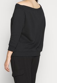 Donna che indossa un top nero a spalle scoperte con maniche 3/4 e pantaloni neri con tasche laterali, in piedi davanti a uno sfondo semplice.