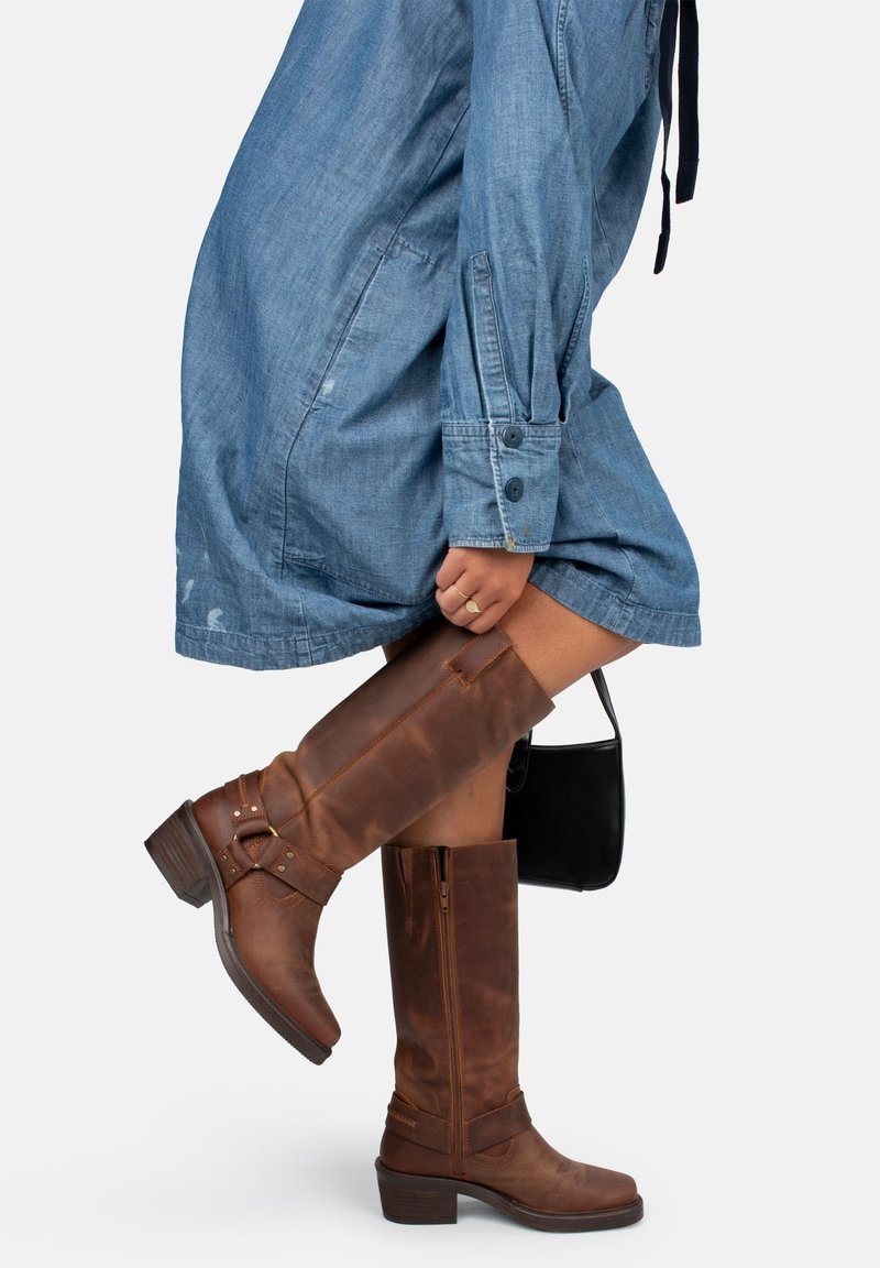 Brown leather knee-high boots with a stacked heel, featuring side zippers, a strap detail, and a smooth texture. Paired with a denim dress.