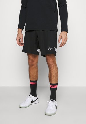 Mann trägt schwarze Nike Sportshorts, schwarze Socken mit pinken Akzenten, weiße Nike Sneaker und ein schwarzes Langarmshirt, steht auf einem grauen Boden.