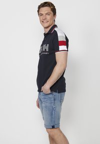 Kék polo ing, kontrasztos piros, szürke és fehér rövid ujjal. Logóval, klasszikus gallérral és oldalsó hasítékokkal rendelkezik, világoskék farmer sorttal párosítva.