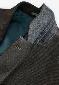 Blazer texturé marron avec col en feutre gris et doublure à motifs verts foncés, mettant en avant un design ajusté et des détails de couture minimaux.