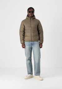 Grüne gepolsterte Jacke mit hohem Kragen, Reißverschluss und gerippter Textur; kombiniert mit hellblauen Jeans und cremefarbenen Sneaker.