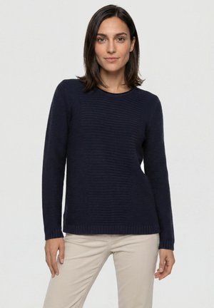 Vrouw met donker haar die een marineblauwe, gestructureerde trui met lange mouwen en beige broek draagt, staand tegen een effen lichte achtergrond.