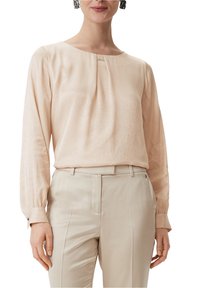 Kvinde iført en lys beige langærmet bluse med plissering, parret med matchende beige høj talje bukser.