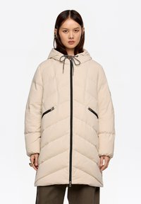 Abrigo acolchado beige con capucha, con un patrón en chevron, cremalleras negras y cierre con cordón; diseñado con bolsillos delanteros y una silueta larga.