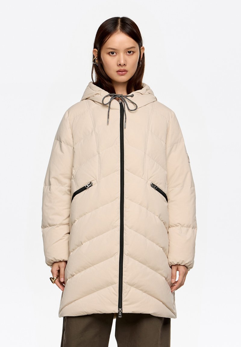 Abrigo acolchado beige con capucha, con un patrón en chevron, cremalleras negras y cierre con cordón; diseñado con bolsillos delanteros y una silueta larga.