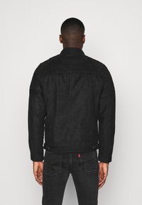 Man som står med ryggen mot kameran, iklädd en svart mockajacka och mörka jeans med synliga bakfickor och en röd etikett.