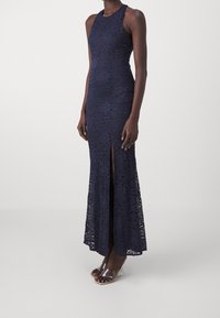 Abito in pizzo blu navy con un design aderente, senza maniche e uno spacco alto fino alla coscia, caratterizzato da intricate fantasie floreali. Sandali con tacco trasparente.