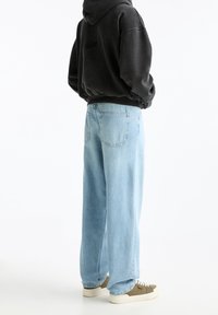 Jeans de mezclilla azul claros con un corte holgado, que cuentan con dos bolsillos traseros y detalles cosidos, combinados con una sudadera negra con capucha y zapatillas deportivas.
