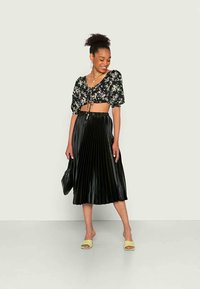Top cropped preto com padrão floral e mangas bufantes, combinado com uma saia midi plissada preta. A modelo usa mules verde claro e segurando uma bolsa preta.